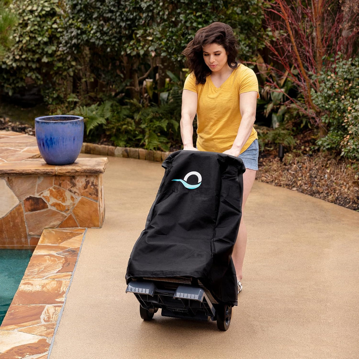 Telo di copertura nero su caddy per robot piscina, spinto da una donna su un vialetto vicino alla piscina.