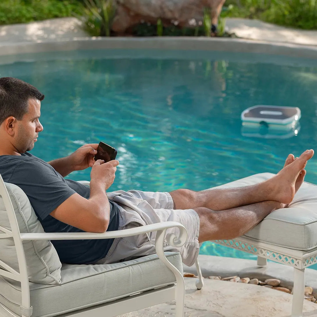 Dolphin Skimmi galleggia su piscina, uomo seduto controlla robot con smartphone, ambiente rilassante.