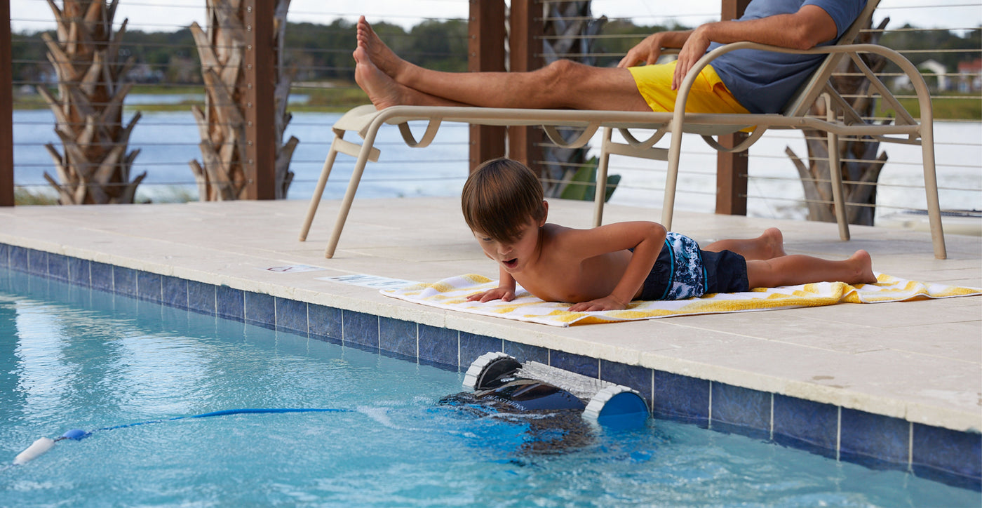 Bambino osserva un robot pulitore in azione nella piscina, mentre un adulto si rilassa su una sdraio.